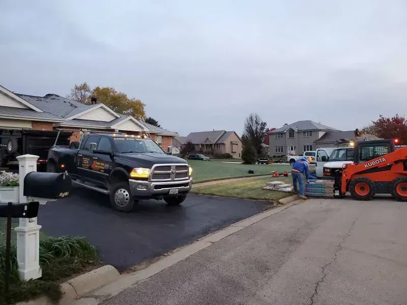 Black pickup truck parked in a suburban driveway beside a driveway crew and orange skid steer machine