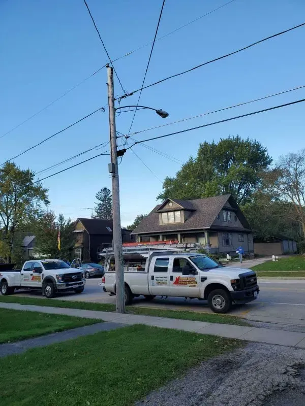 Two utility trucks parked on a residential street beside a wooden house and utility pole.