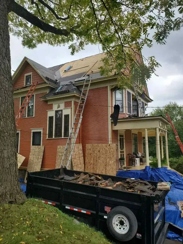 Damaged red house with plywood-covered windows, roof work in progress, and a debris trailer in front