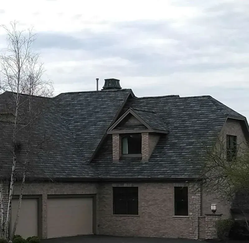 Gray brick house with dark shingled roof, garage, and dormer windows under a cloudy sky