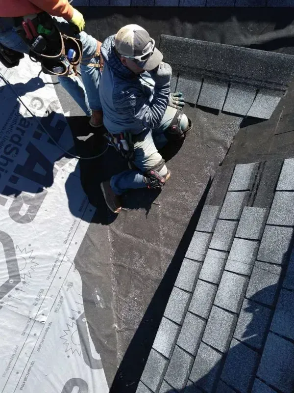 Roofers installing shingles on a steep roof beside a white tarp-covered section.