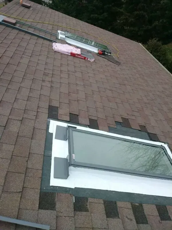 Sloped shingle roof with a skylight and rooftop materials near the ridge, overlooking trees.