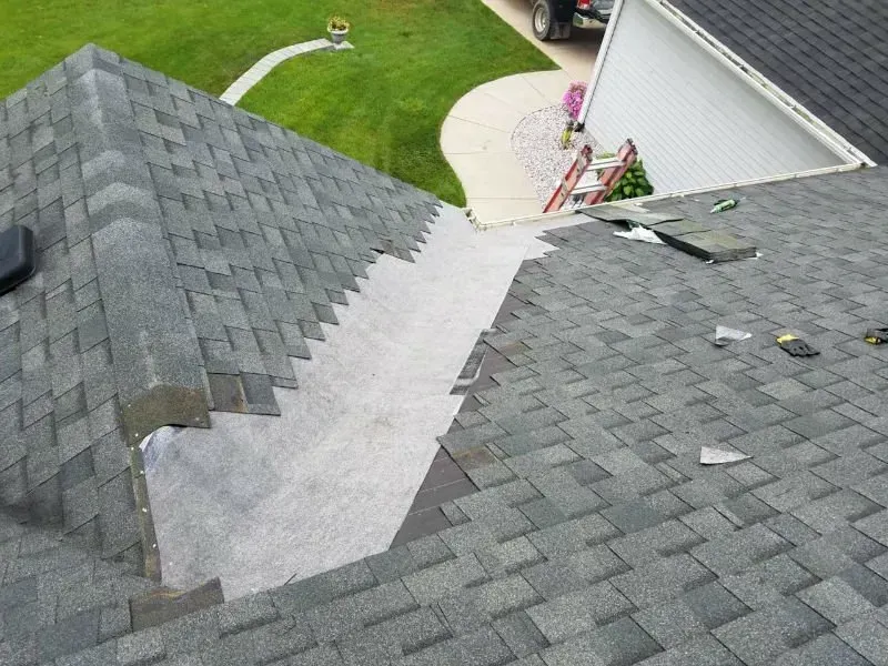 Roof under repair with gray shingles and a tarp-covered section beside a driveway and lawn