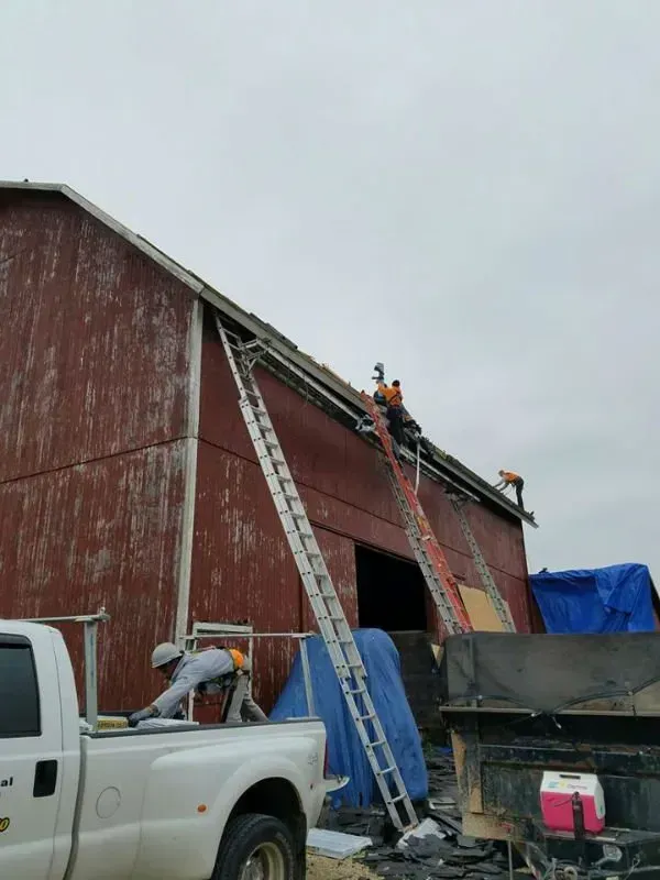 Workers on a barn roof with ladders and a pickup truck in a rural worksite