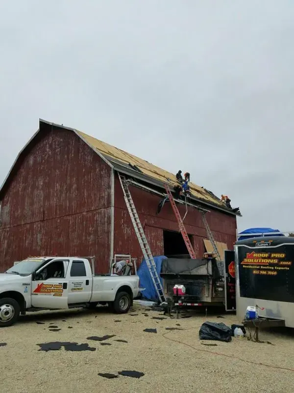 Workers on a red barn roof, with a ladder, white pickup truck, and work van in a gravel lot.