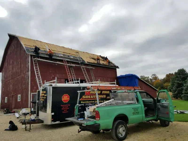 Workers repairing a red barn roof beside a green work truck and trailer under a cloudy sky
