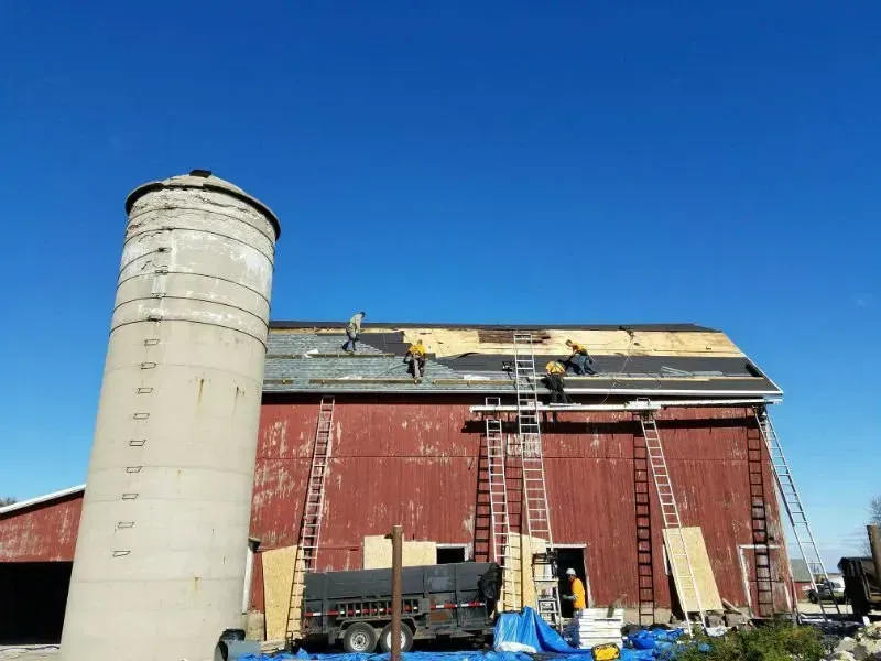 Red barn with damaged roof beside a white silo under a clear blue sky.