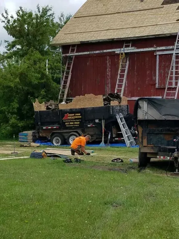 Worker in orange vest near a red barn, with trucks and ladders in a grassy yard.