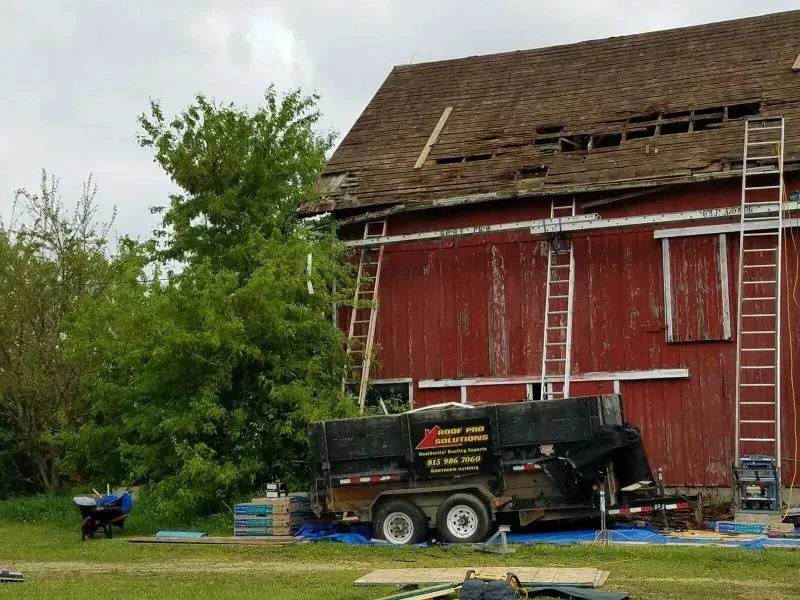 Red barn under repair with ladder, trailer, and trees in a rural yard