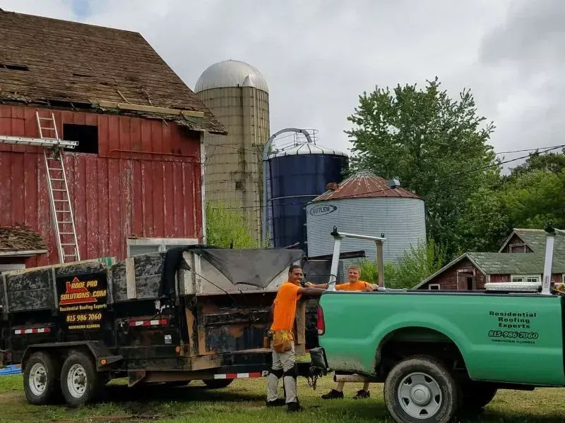 Worker in orange vest beside a green truck and farm trailer near red barn and silos