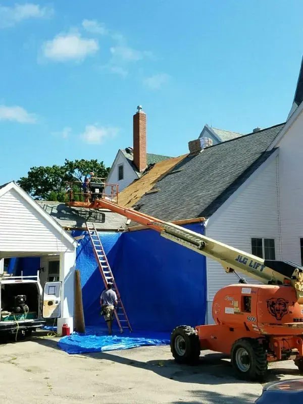 Workers on ladders and a lift repairing a blue tarp-covered roof beside a house and garage.