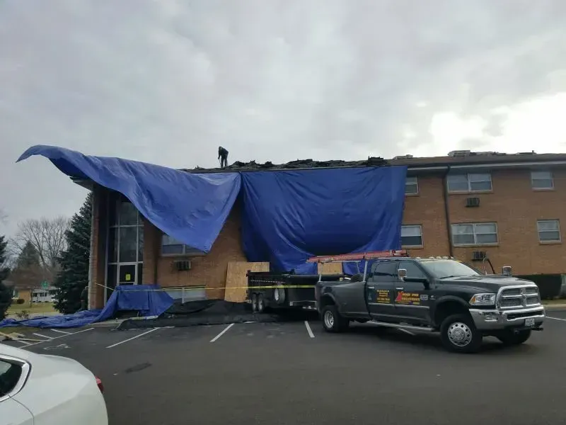 Blue tarp draped over a damaged building with a pickup truck parked in front under an overcast sky