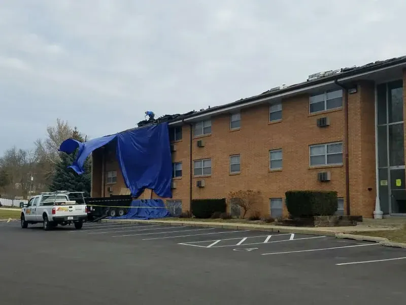 Blue tarp covering damaged roof on brick apartment building, with a pickup truck parked in the lot.