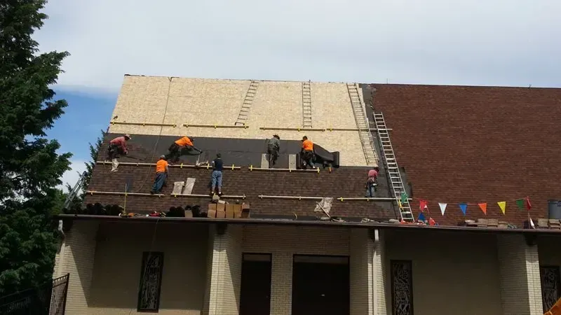 Workers on a roof installing shingles on a house, with ladders and safety gear visible.