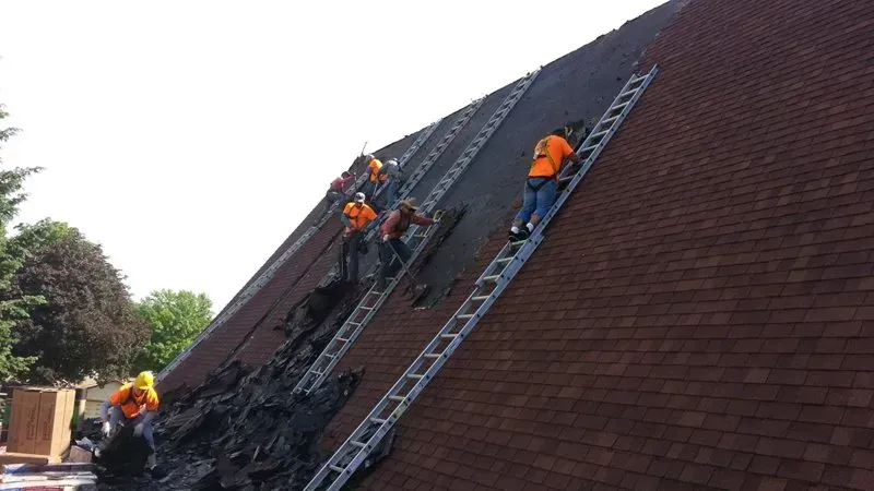 Workers in orange gear climbing ladders and repairing a steep shingled roof