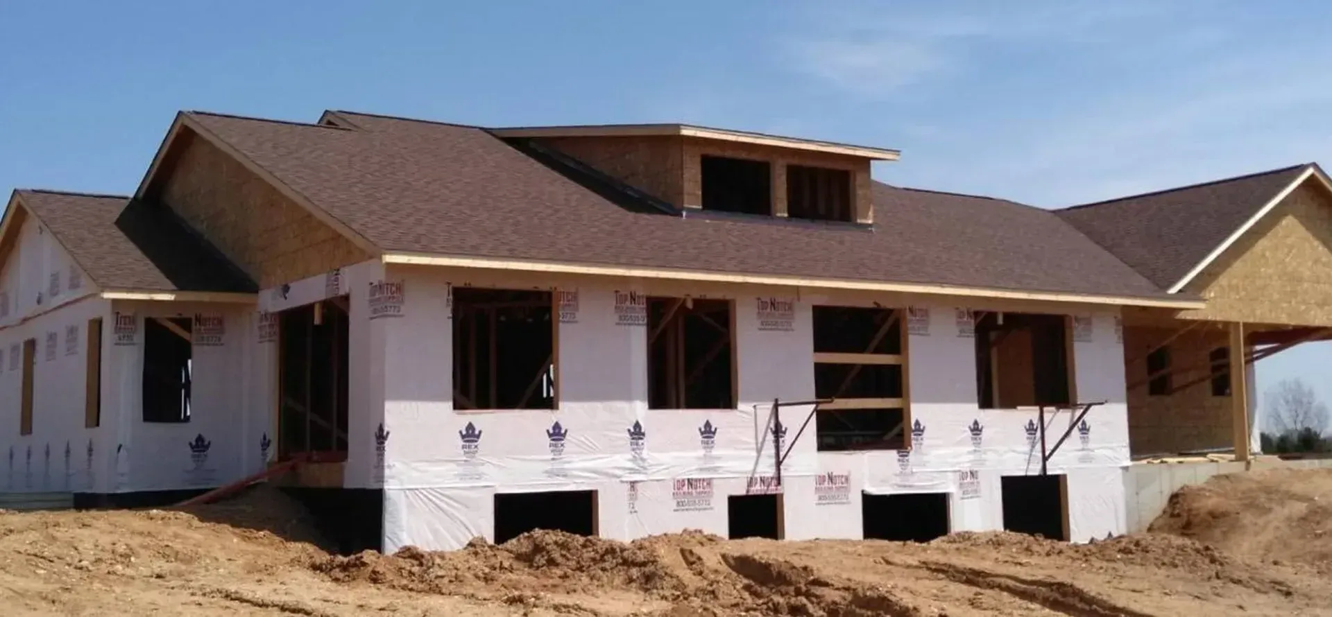 Partially built house with white siding, brown roof, and exposed foundation on a dirt lot
