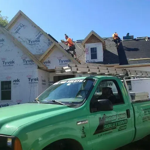 Green roofing truck parked in front of a house under construction, with workers on the roof.