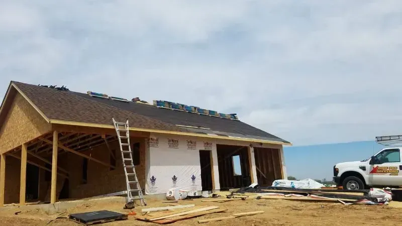Single-story house under construction with ladder, workers, and a pickup truck on a dirt lot.