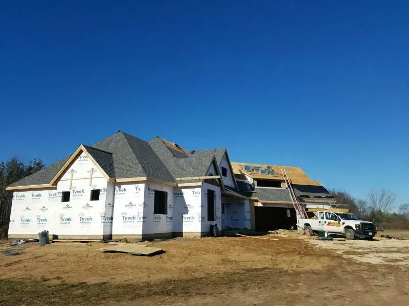 New house under construction with white wrap, gray roof, and a truck parked beside it under a clear blue sky