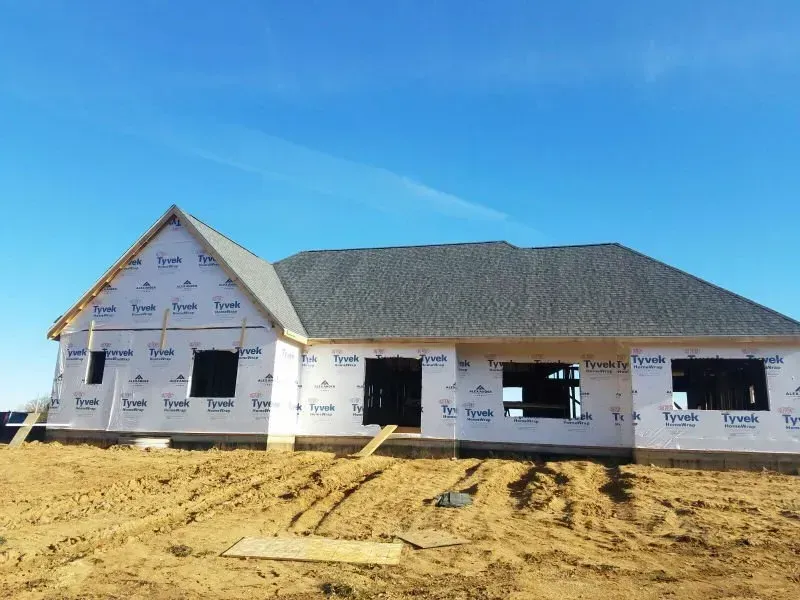 Partially built house with white house wrap and dark roof on a dirt lot under a blue sky