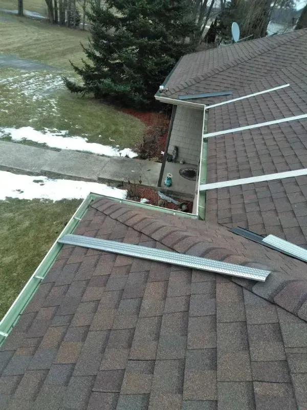 Snow-dusted residential roofs with several long metal planks laid across them near a tree-lined yard