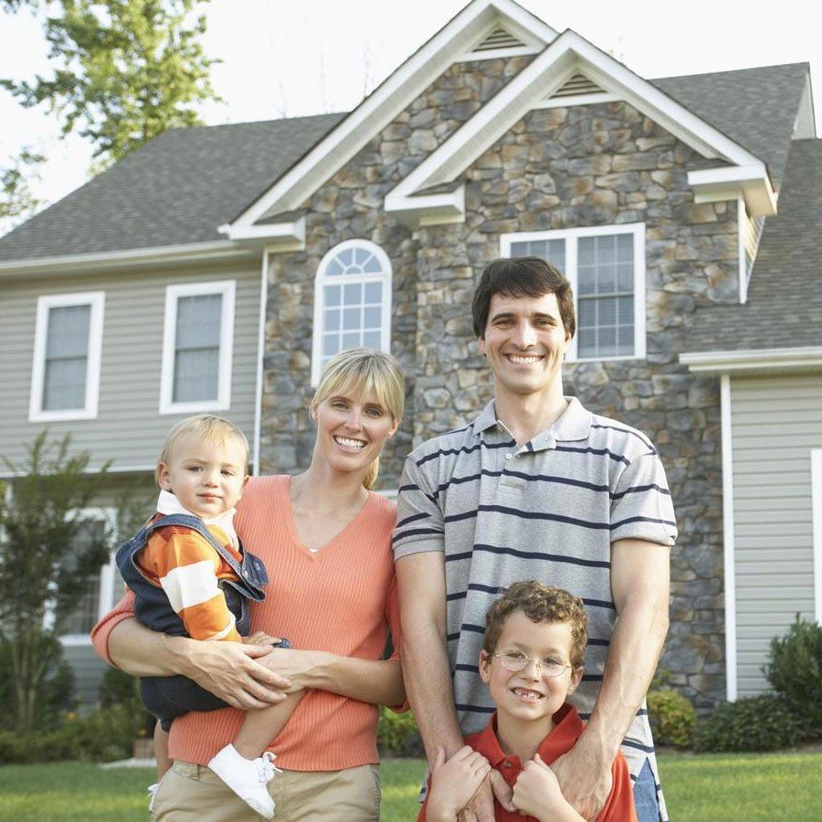 Family of four smiling outside a stone-and-gray house, with two adults holding two young children.
