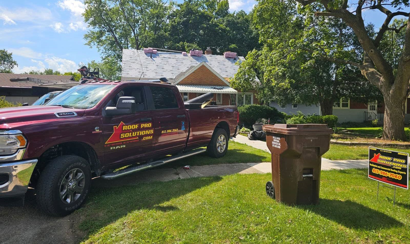 Maroon truck with company sign parked on a sunny lawn beside a brown trash bin and yard sign.