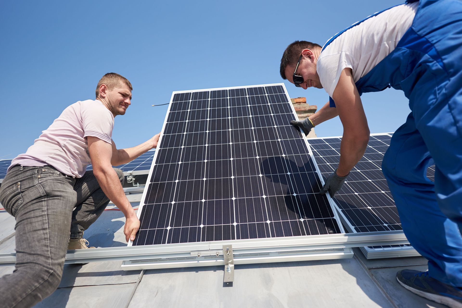 Two workers install solar panels on a rooftop under a clear blue sky.