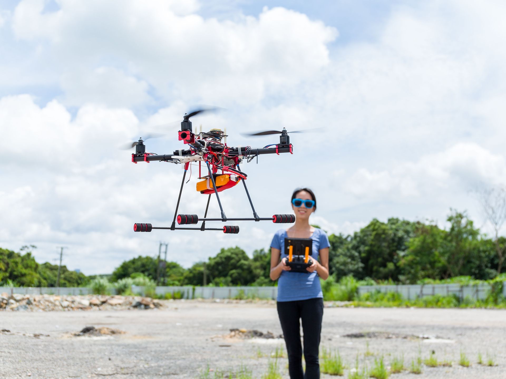 Woman holding a controller beside a large drone in a field under a cloudy sky
