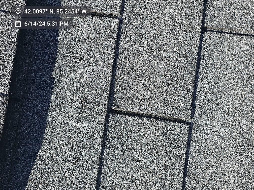 A close-up of dark grey, textured asphalt roof shingles with a vertical shadow cast across the surface.