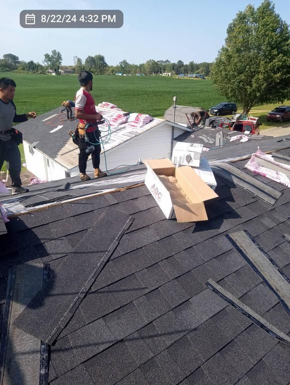 Two workers installing black shingles on a residential roof on a sunny day with a rural field in the background.