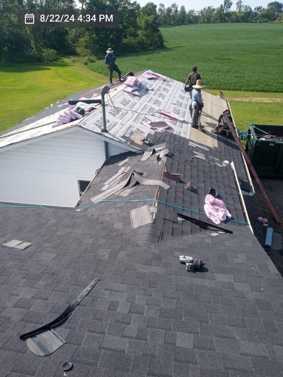 Three workers perform roof repairs on a house, installing shingles and underlayment near a grassy field and dumpster.