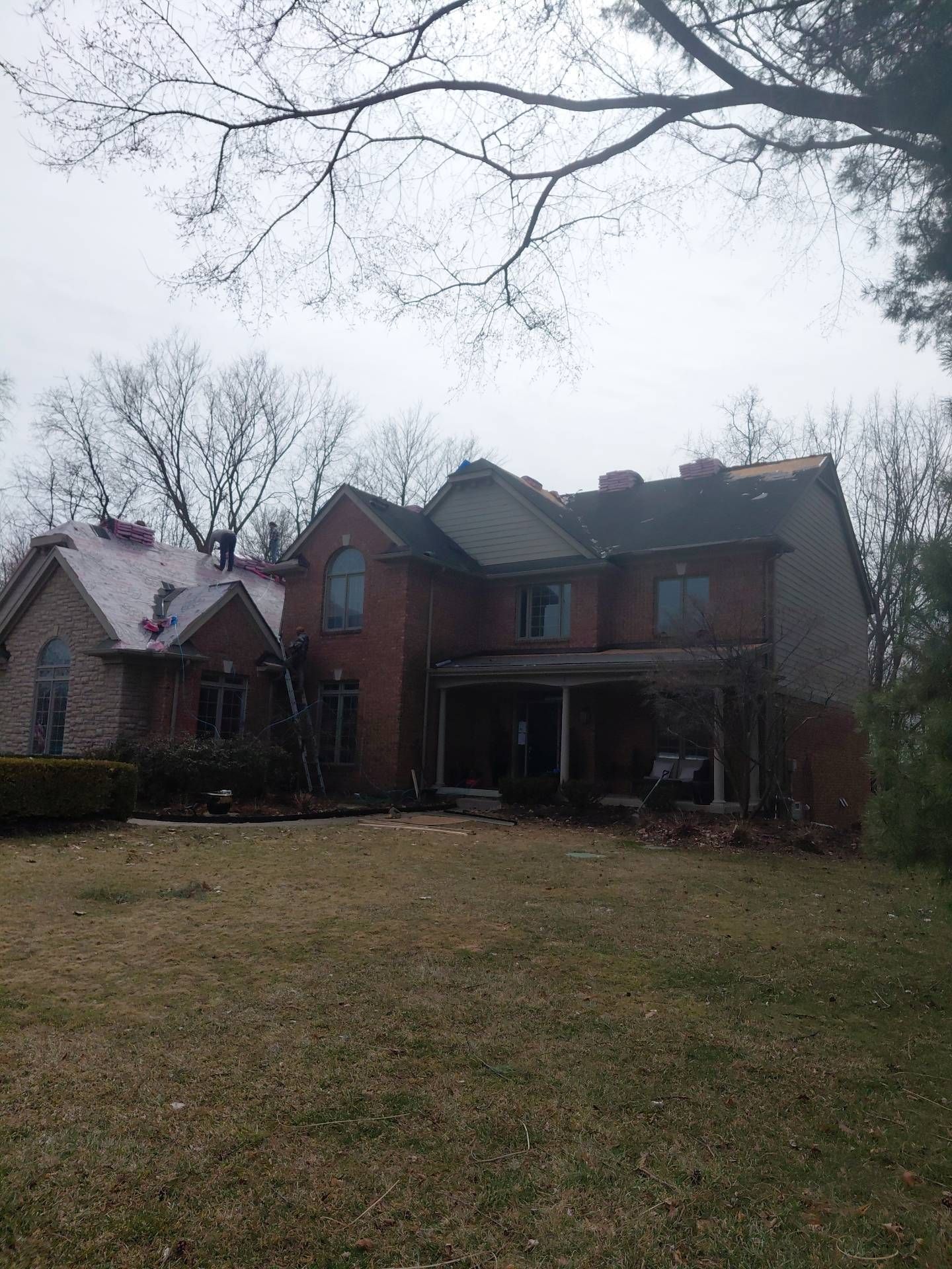 A two-story brick house with a partially replaced grey shingled roof under a cloudy sky.