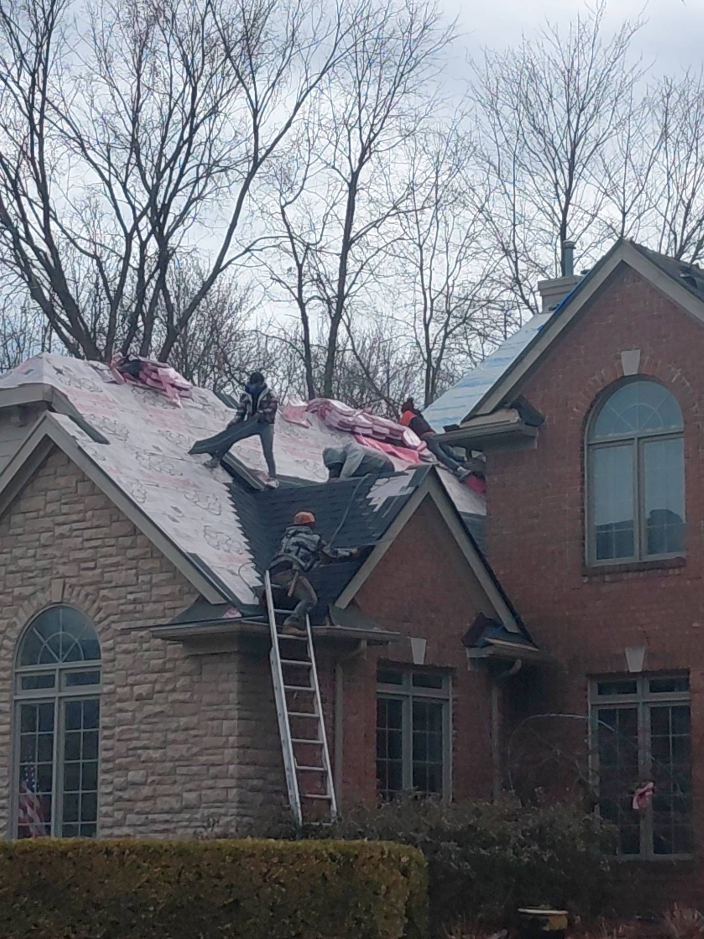 Three workers remove old shingles from the gabled roof of a two-story brick and stone house, using a ladder for access.