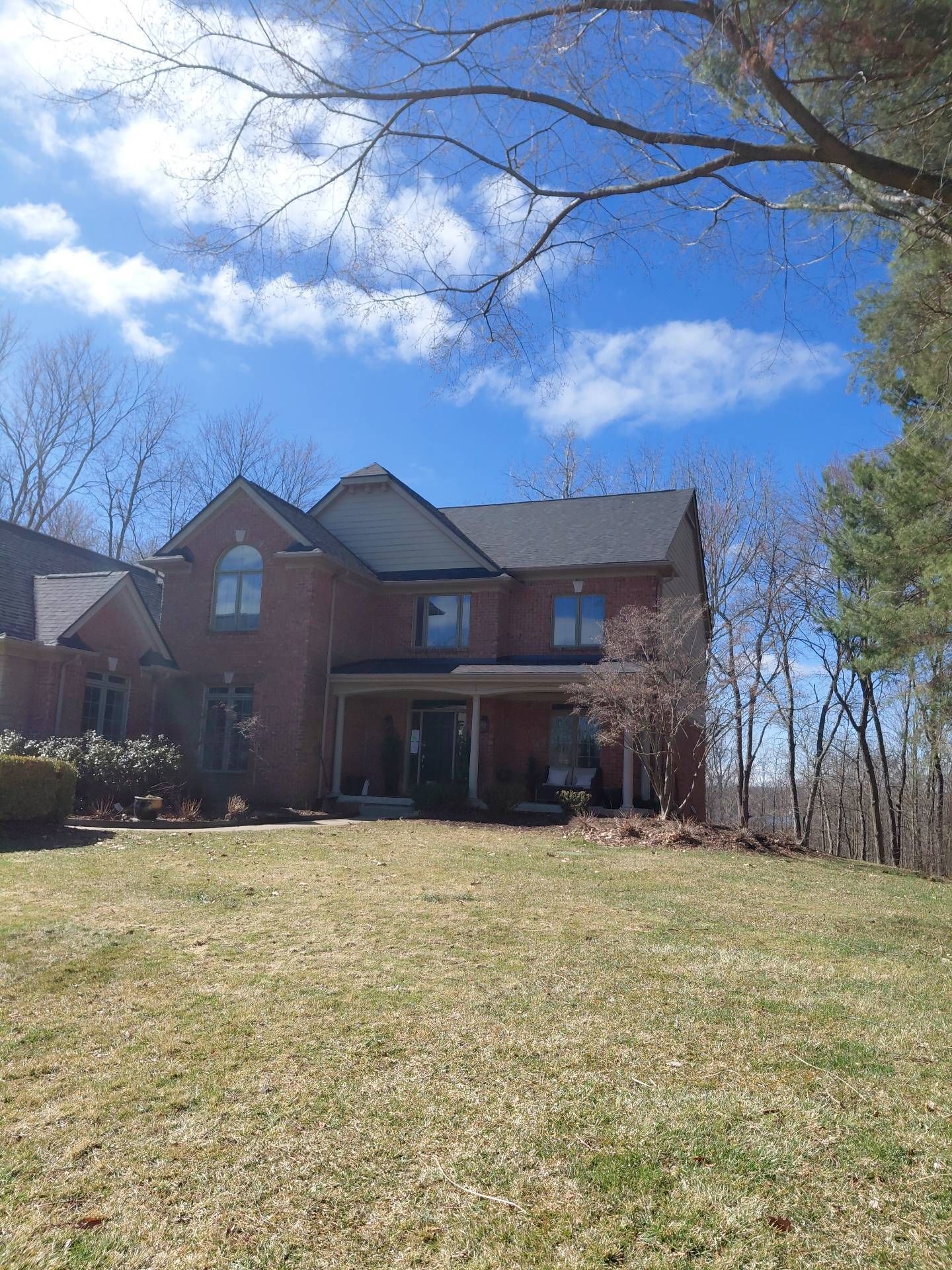 A multi-story brick house with a dark shingled roof stands on a grassy lawn under a clear blue sky with thin trees.