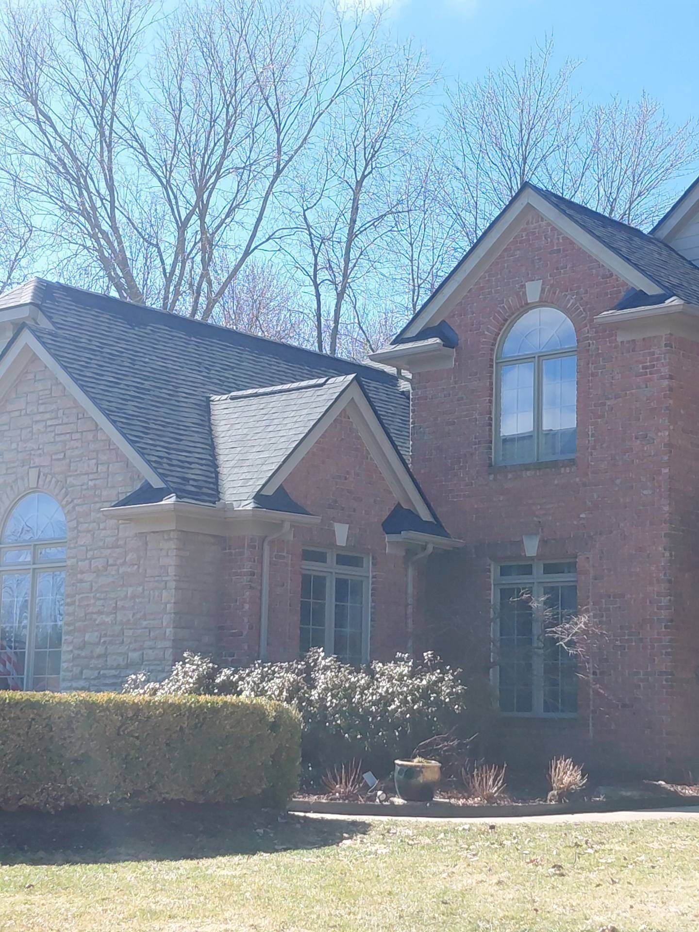 A two-story brick house with a stone facade section, grey roof shingles, and arched windows under a clear blue sky.