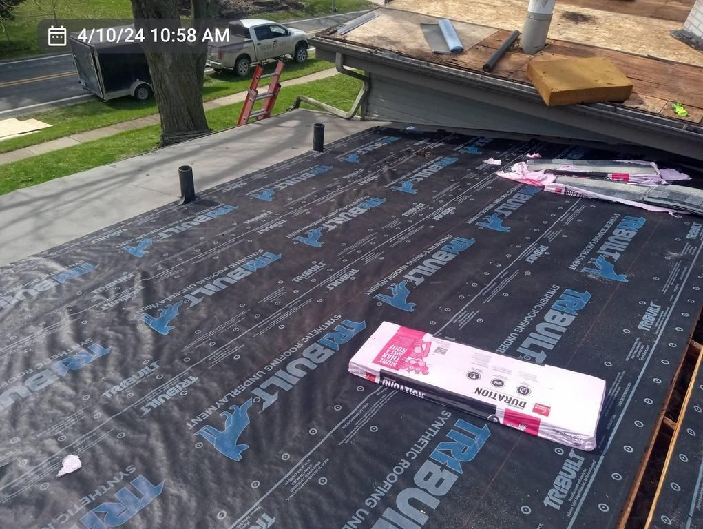 A roof under construction covered in black synthetic underlayment, with a box of roofing shingles and plumbing vents.