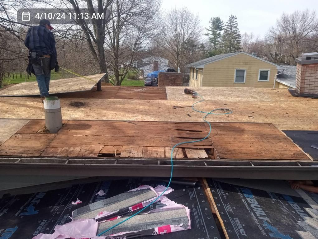 A person working on a house roof during a replacement project, with exposed plywood decking and roofing materials below.
