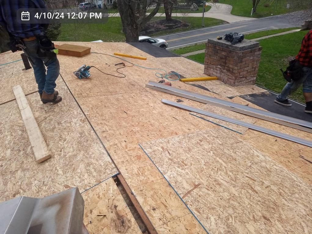 Two people work on a residential roof replacement, installing plywood sheathing near a brick chimney.