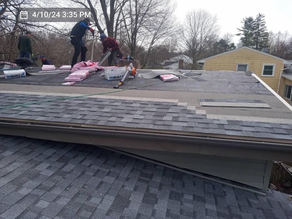 Workers install shingles on a residential roof, with bundles of roofing materials visible on the flat roof section.