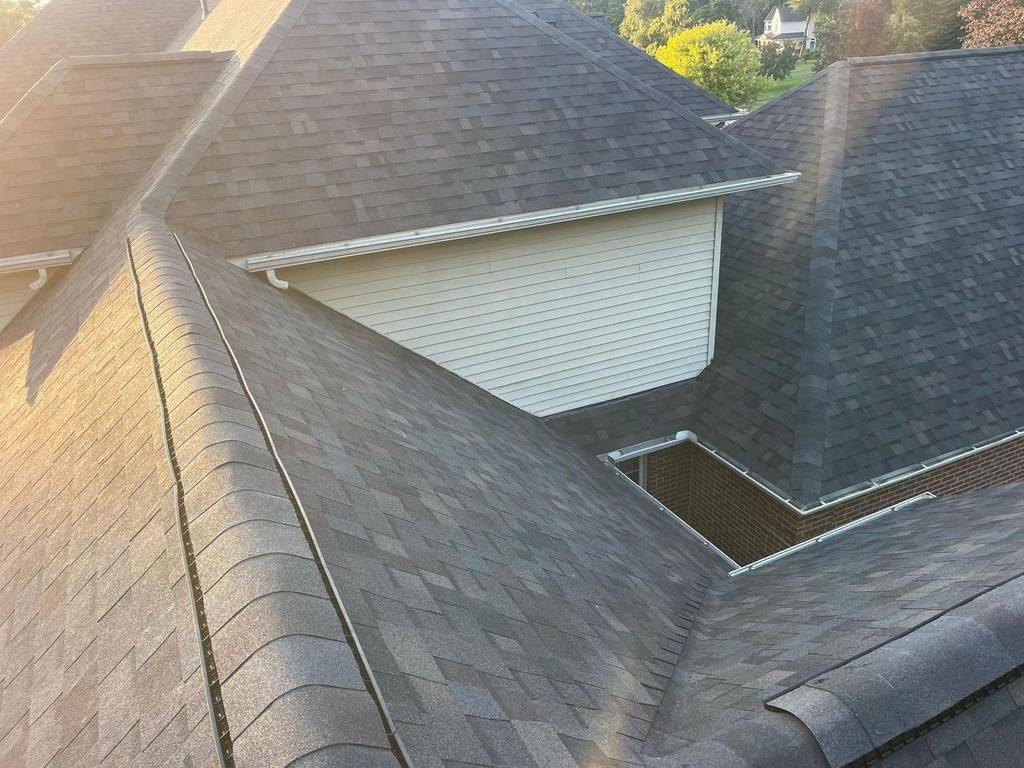 A high-angle view of gray asphalt shingled roofing sections, including a white-sided dormer and built-in gutters.