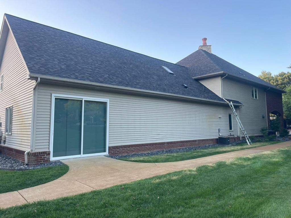 A tan-sided house with a dark shingled roof, a sliding glass door, and a brick base, viewed from a grassy lawn.
