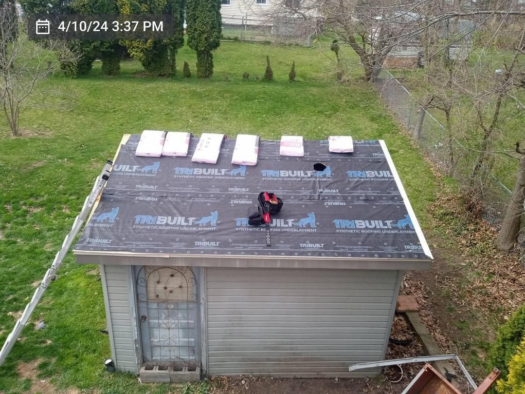A shed roof covered in black underlayment with several stacks of shingles laid out on top and a ladder leaning against it.