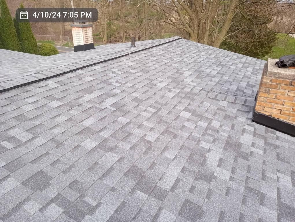 A close-up view of a recently installed gray, patterned asphalt shingle roof with a brick chimney in the background.
