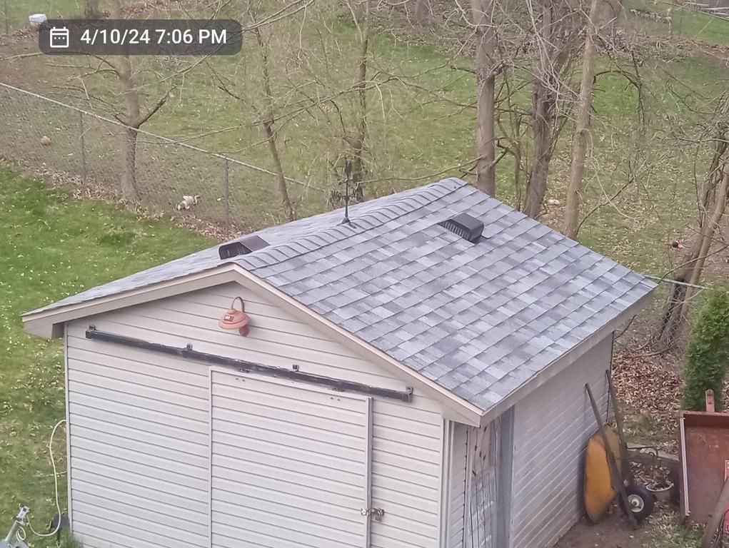 A high-angle view of a backyard shed with a grey shingled roof, sliding door, and vents, set in a grassy yard.