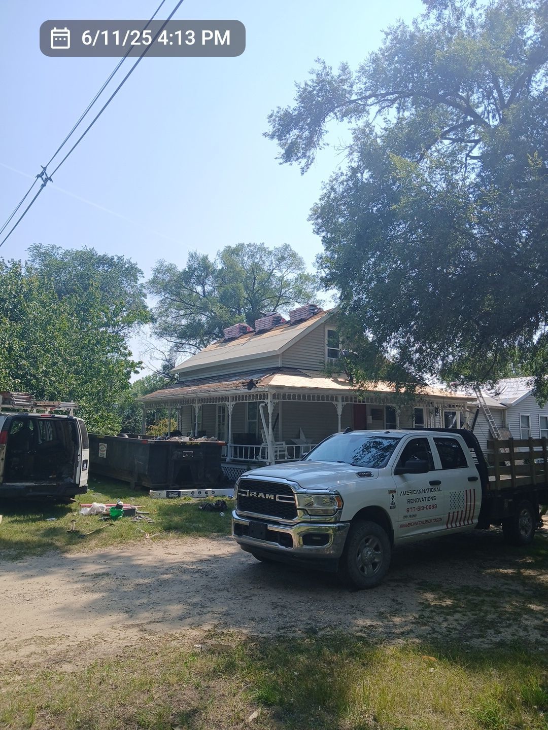 A white Ram work truck parked in front of a residential house undergoing roof repairs, with a dumpster and trees nearby.