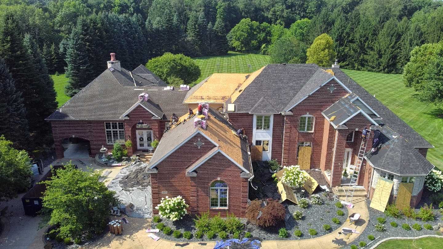 An aerial view of a red brick house under construction, showing partially replaced roof shingles and exposed wood decking.
