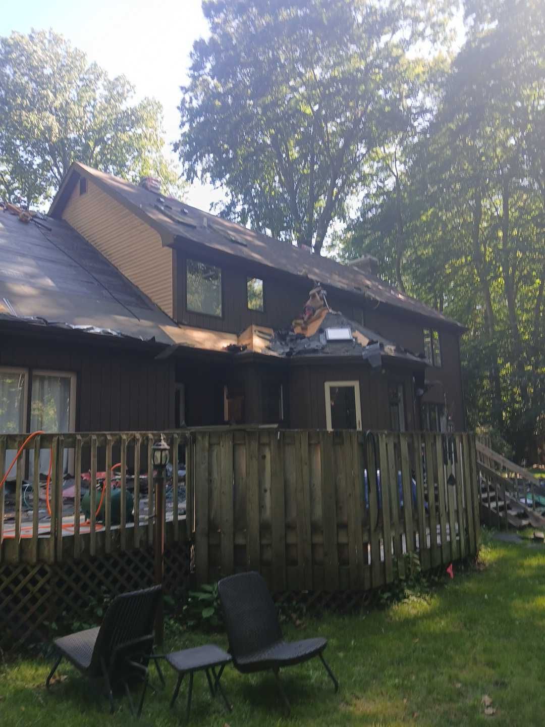 A two-story dark brown house with an attached wooden deck, surrounded by trees on a sunny day.