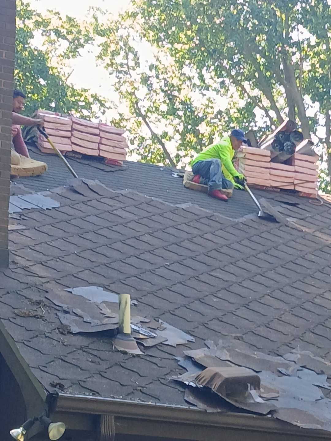 Two workers in bright shirts remove shingles from a residential roof while standing near stacks of new roofing material.