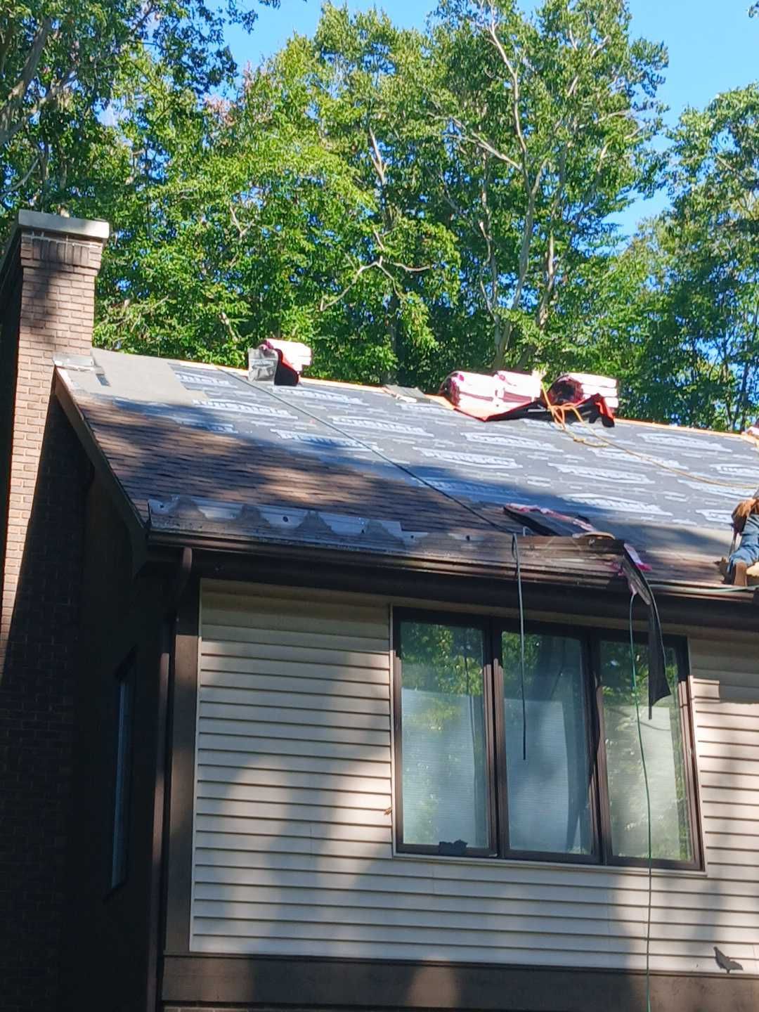 A house roof under construction, featuring grey underlayment, bundles of shingles, and a chimney against a tree background.